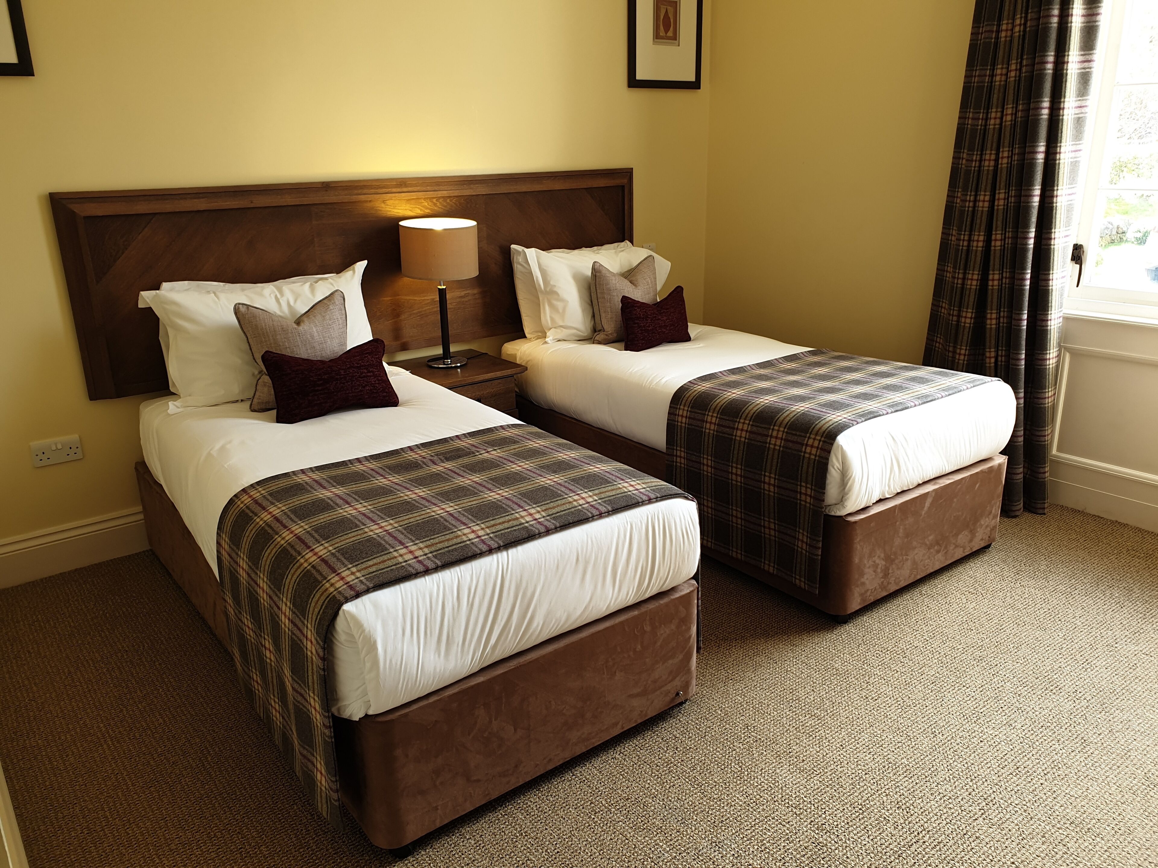 A hotel room with two beds, a nightstand, a lamp, and a window with curtains.