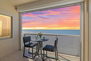 A balcony with a view of the ocean at sunset, featuring two wicker chairs and a small table with a vase of flowers. - Brand New Rental - Beachfront 2BR with Gulf Views, Heated Pool & In-Unit Laundry (Fort Myers Beach)