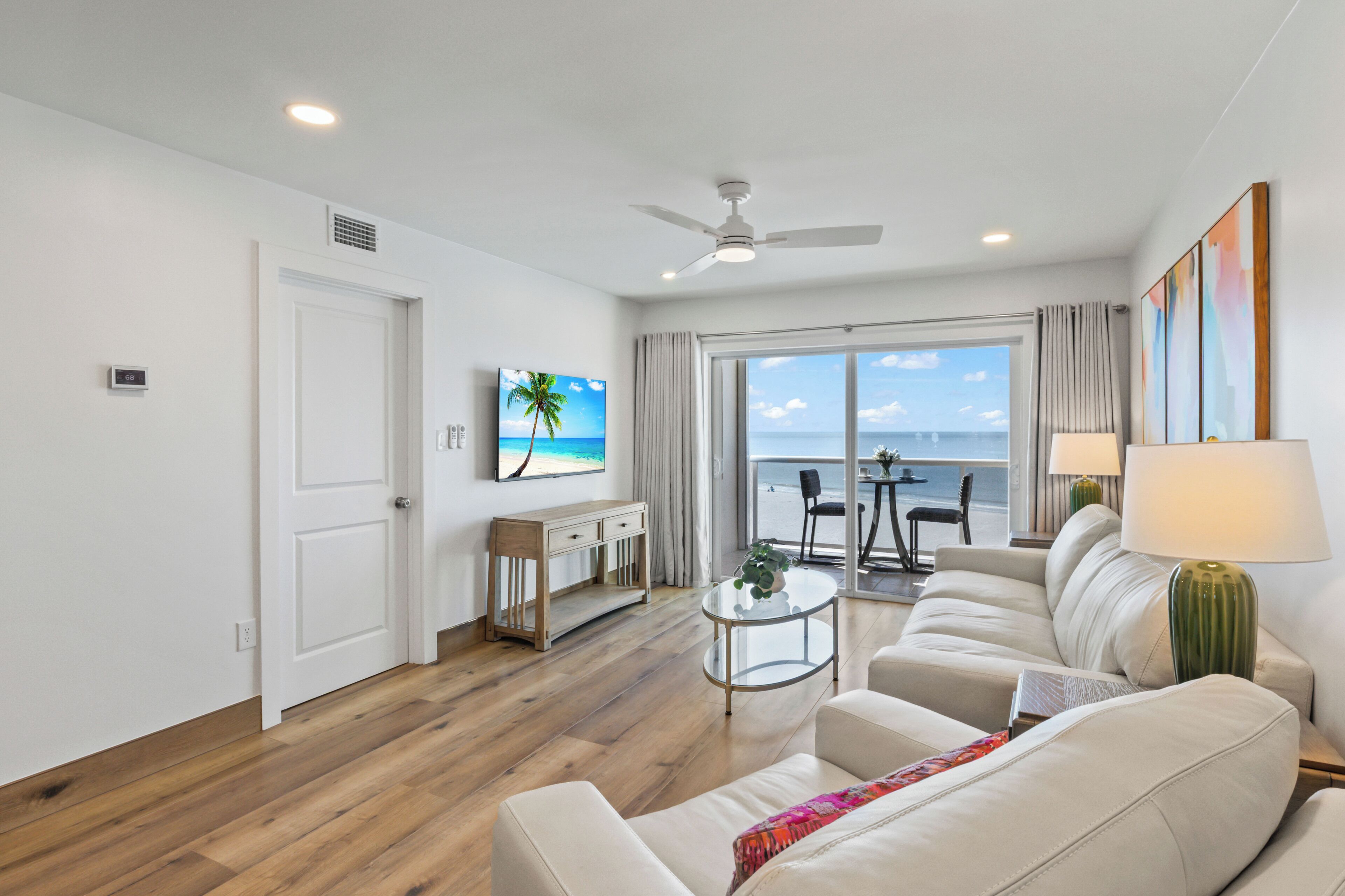 A modern living room with a sofa, a coffee table, and a TV on the wall. There is a window with a view of the ocean.