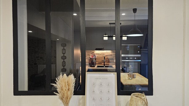 A modern kitchen with a large window, a framed zodiac signs poster, and a vase of pampas grass on a wooden table. - La Charmante (Puyoô)