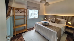 A bedroom with a bunk bed, a window with a view of buildings, a bedside table with a lamp, and a hanging light fixture.
