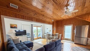 A wooden-paneled room with a sectional sofa, a flat-screen TV, a dining area with chairs and a table, and a refrigerator. - Sea Esta: Perfect Getaway on the Canal with Boat Dock (Ocracoke)