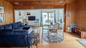 A cozy living room with a dark blue sofa, a wooden dining table with chairs, a television, and a view of the outside. - Sea Esta: Perfect Getaway on the Canal with Boat Dock (Ocracoke)