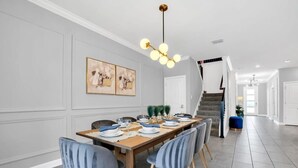 Un coin repas est aménagé avec une longue table en bois, dressée pour six personnes, et entourée de six chaises aux pieds en bois. Un lustre est suspendu au-dessus de la table, et les murs sont peints dans une teinte de gris clair.