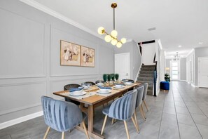 Ein Essbereich mit einem langen Holztisch, gedeckt für sechs Personen, umgeben von sechs Stühlen mit Holzbeinen. Über dem Tisch hängt eine Chandelier, und die Wände sind in einem hellgrauen Farbton gestrichen.