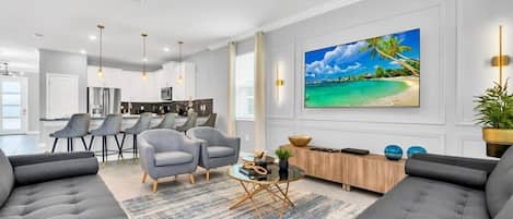 Una sala de estar moderna con sofás grises, una mesa de centro y un televisor grande que muestra una escena de playa.