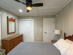 Quarto com cama, penteadeira, guarda-roupa, ventoinha no teto e mesa de cabeceira com abajur.