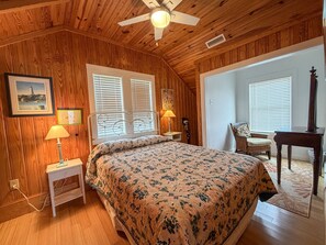 A bedroom with a wooden ceiling, a bed with a floral bedspread, two bedside lamps, a chair, a table, and a picture on the wall. - Sea-Esta Upstairs: A Couple's Getaway on the Canal (Ocracoke)