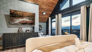 Un dormitorio con una cama grande, vistas a las montañas a través de las ventanas, una chimenea y un cuadro en la pared.