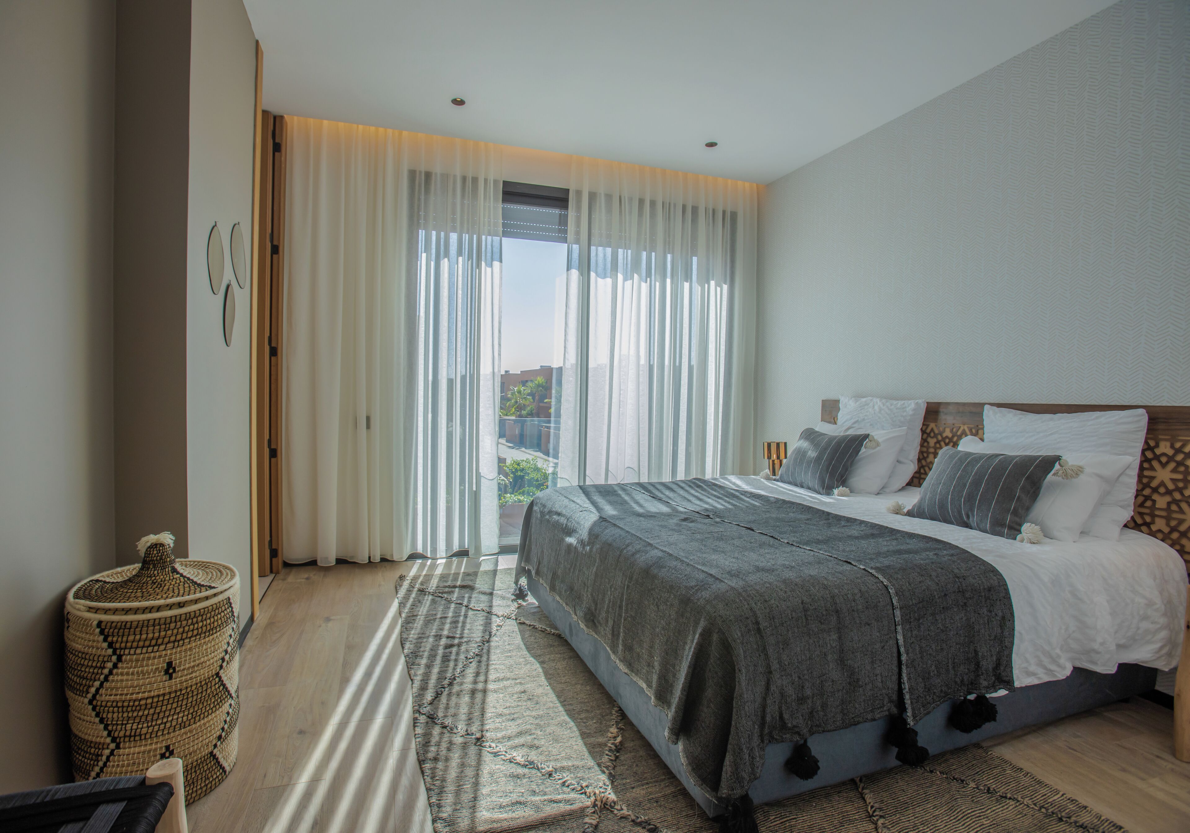 A modern bedroom with a large bed, a view through sheer curtains, and a woven floor mat.