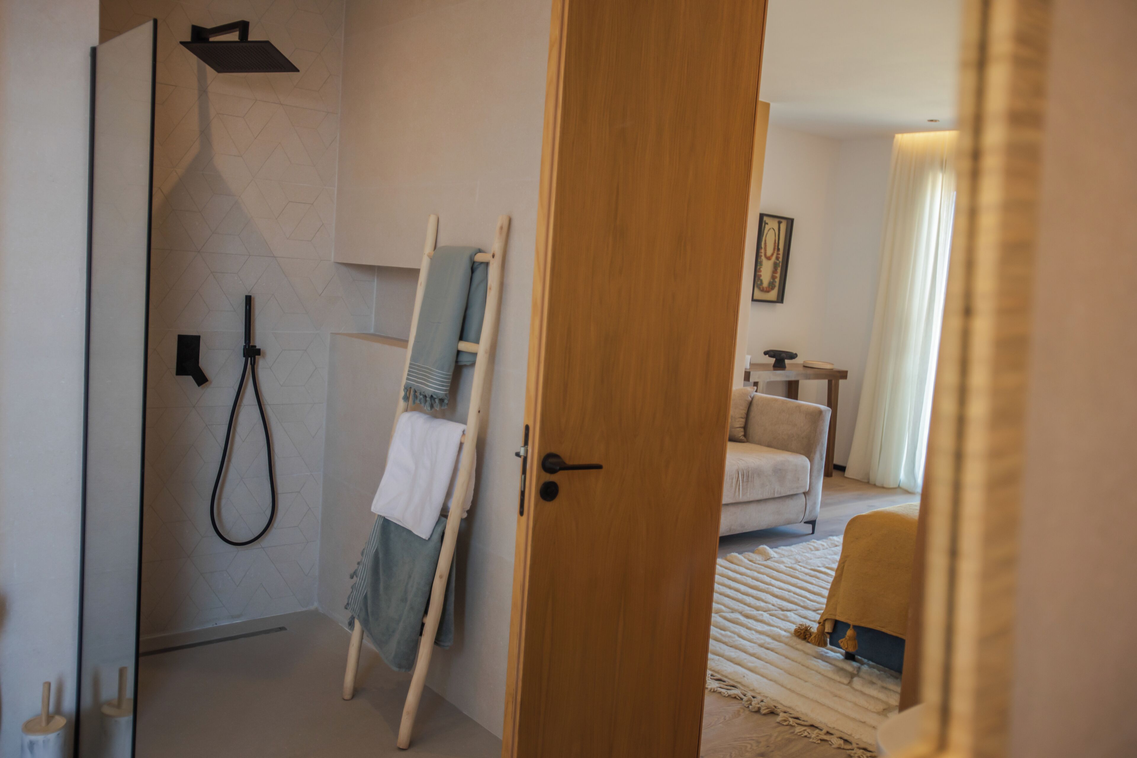 A hotel room with a wooden door, a shower area with a towel rack, a bed with a white bedspread, and a small table with a lamp.