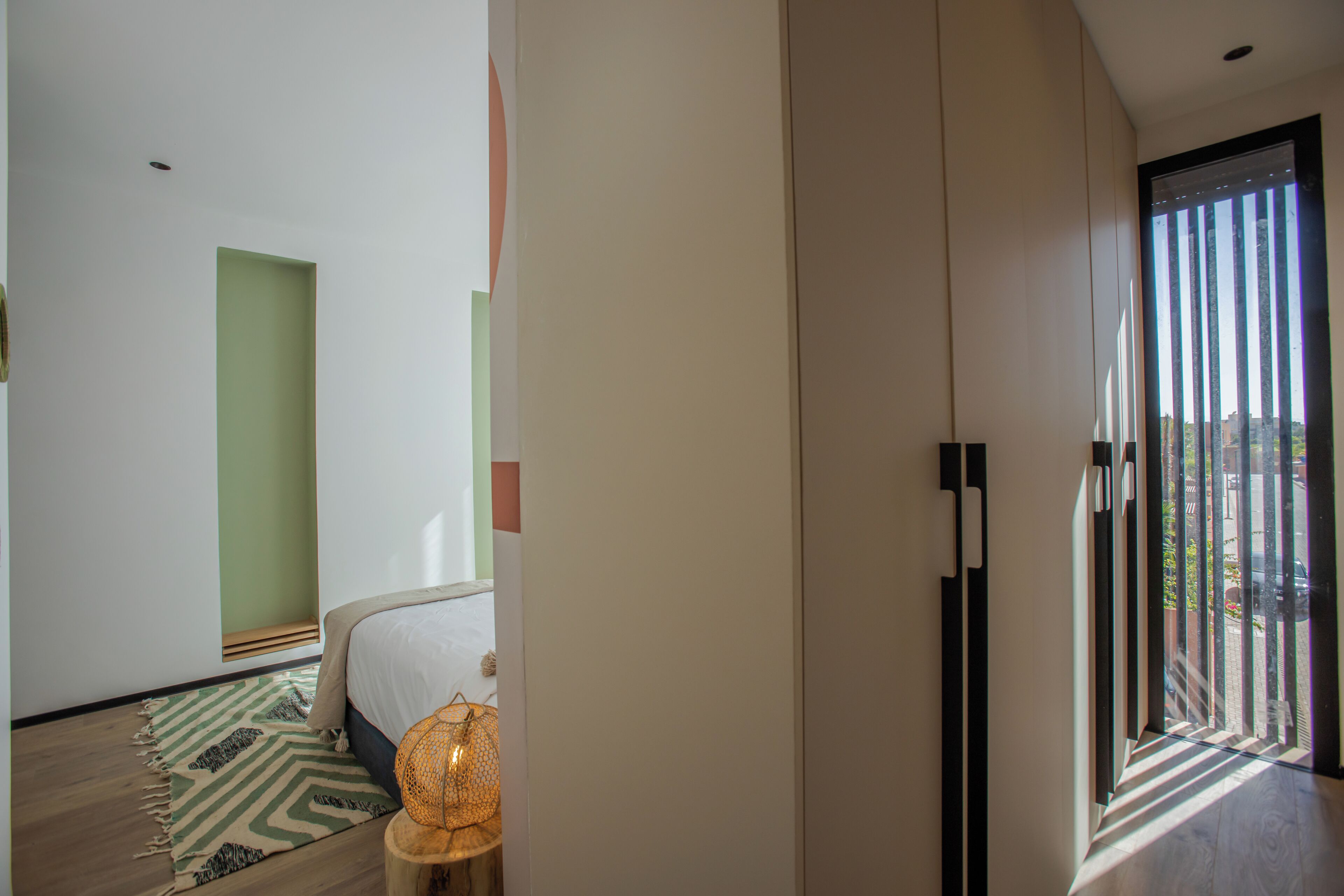 A modern hotel room with a bed, a green accent wall, a patterned rug, and a view through a window.