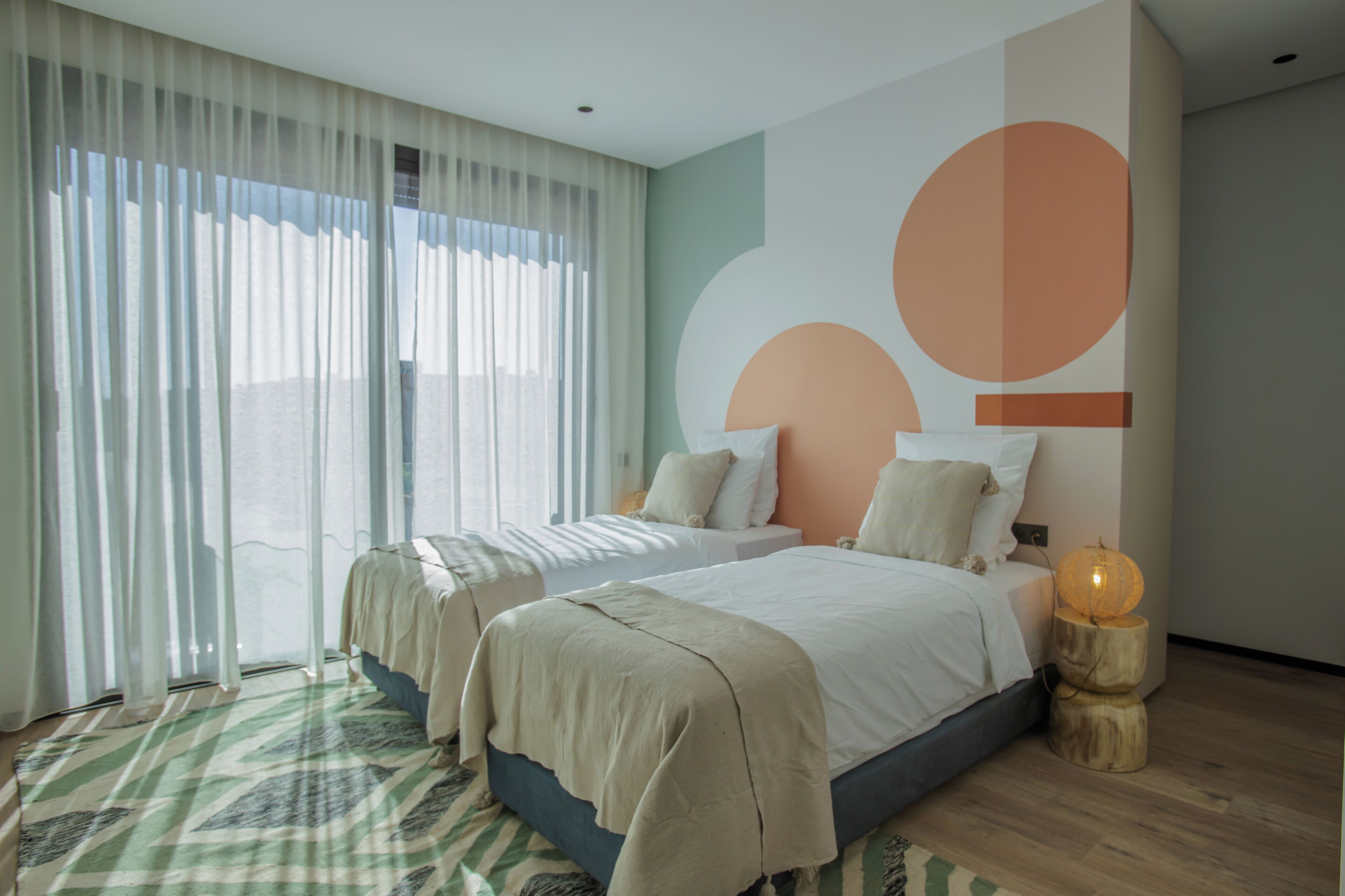 Een moderne hotelkamer met twee bedden, een groot raam met dunne gordijnen, een geometrisch wandontwerp en een nachtkastje van hout met een lamp.