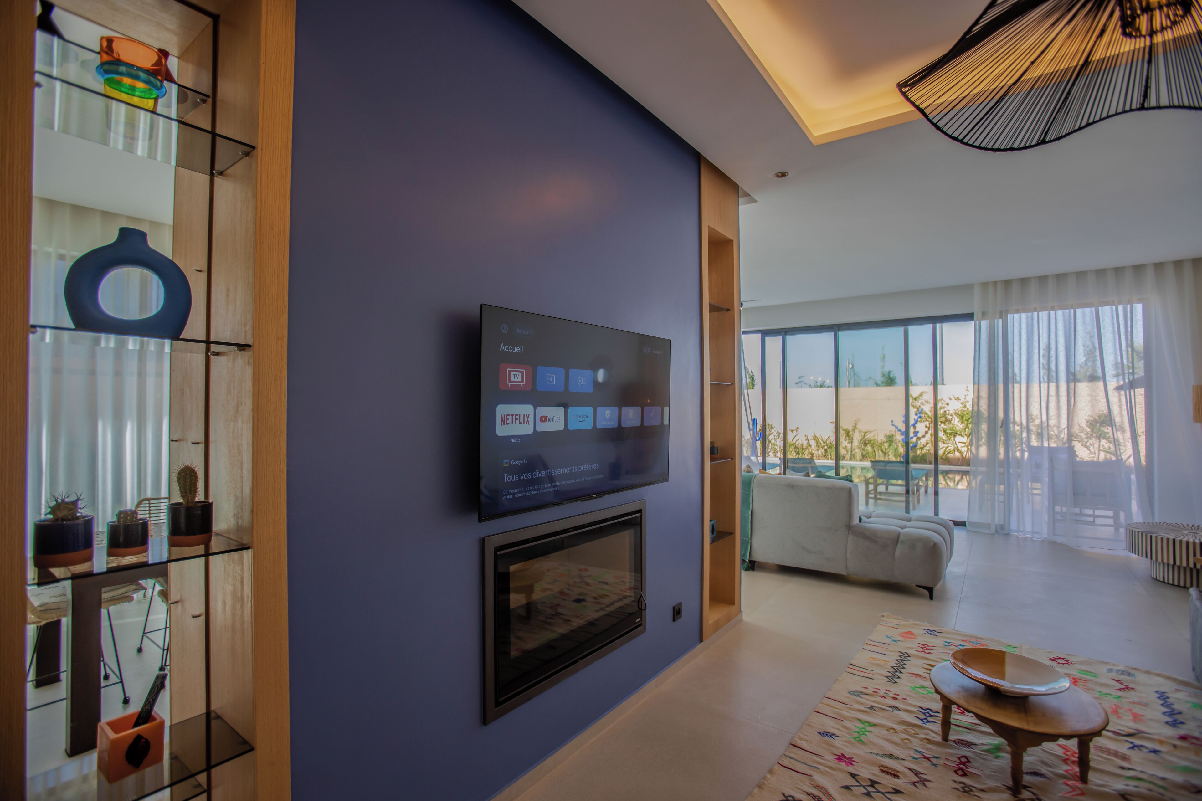 A modern living room with a flat-screen TV mounted on the wall, a glass shelving unit with decorative items, a sofa, and a large window offering a view of the outdoors.