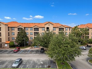 A multi-story building with a red-tiled roof, parking lot, and green landscaping. - Spacious 3-bedroom condo Just Mins from Disney! (Kissimmee)