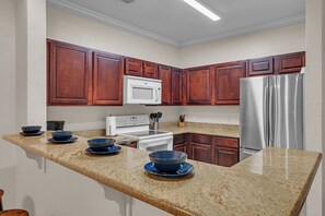 A kitchen with granite countertops, wooden cabinets, a stainless steel refrigerator, and a white microwave. - Spacious 3-bedroom condo Just Mins from Disney! (Kissimmee)
