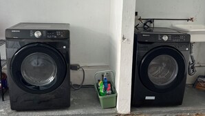 Zwei schwarze Samsung Waschmaschinen in einem Waschraum mit einem weißen Waschbecken und einem grünen Korb voller Reinigungsmittel.