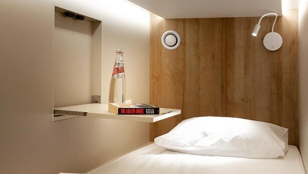 A modern hotel room with a wooden headboard, a bedside table with a bottle and a book, and a white bed with a rolled towel. - Jeru Caps CityCenter (Jerusalem)