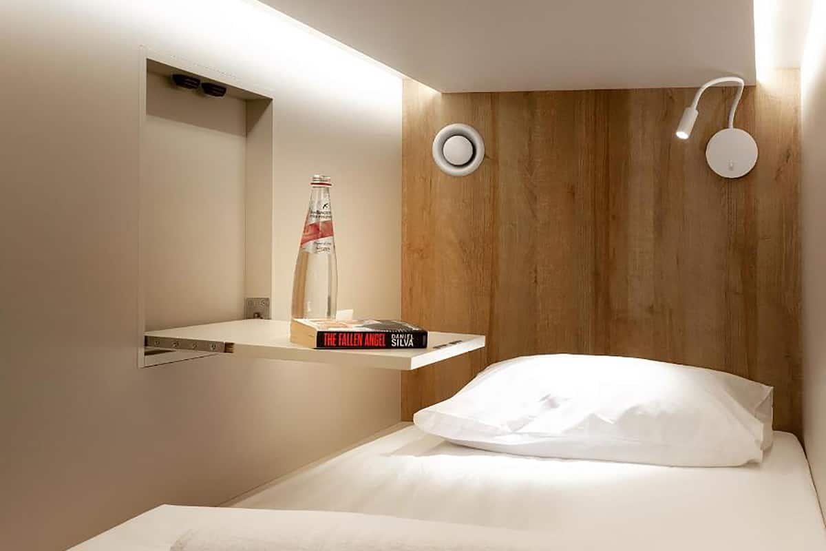 Habitación de hotel moderna con cabecera de madera, una mesita de noche con una botella y un libro, y una cama blanca con una toalla enrollada.