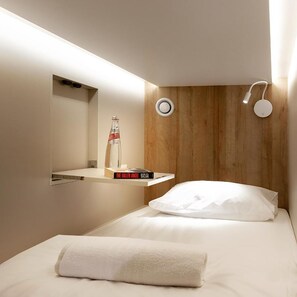 A modern hotel room with a wooden headboard, a bedside table with a bottle and a book, and a white bed with a rolled towel. - Jeru Caps CityCenter (Jerusalem)