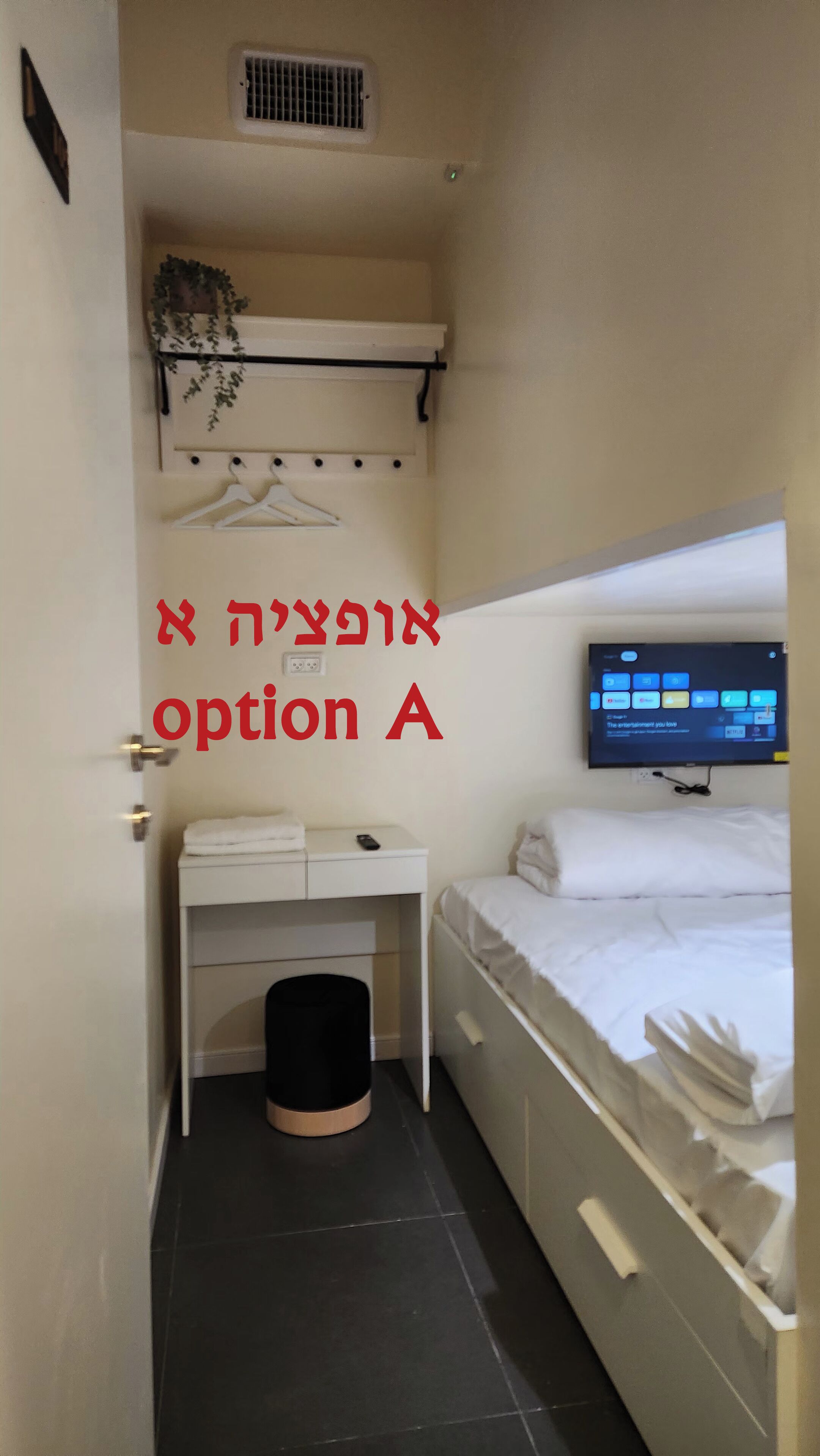 A small, compact hotel room with a bed, a desk, and a TV mounted on the wall.