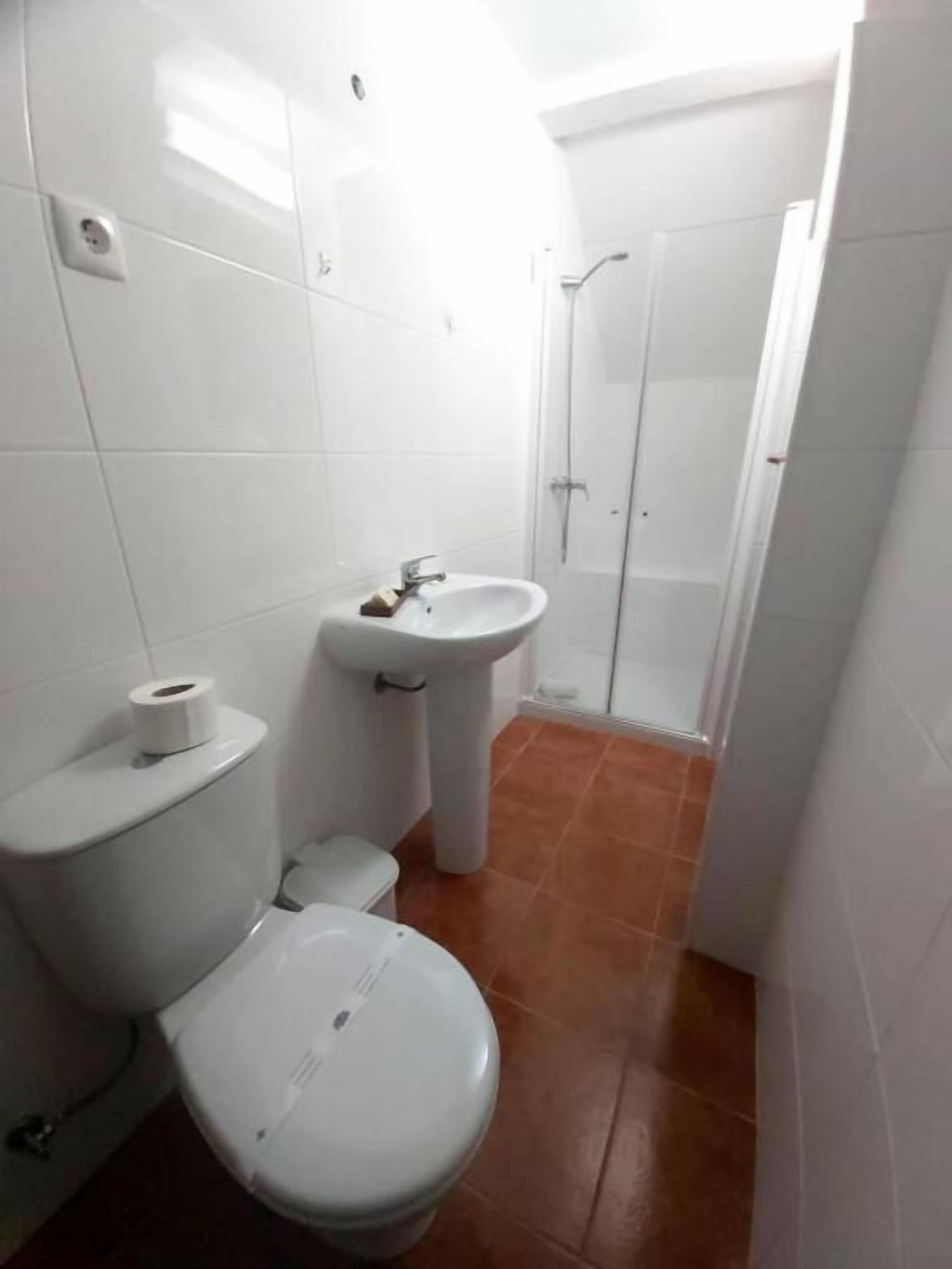 A small, compact bathroom with white tiles, a toilet, a sink, and a shower.