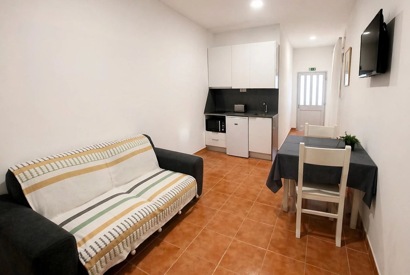 A compact living space with a sofa, a dining table with chairs, a kitchenette, and a flat-screen TV mounted on the wall.