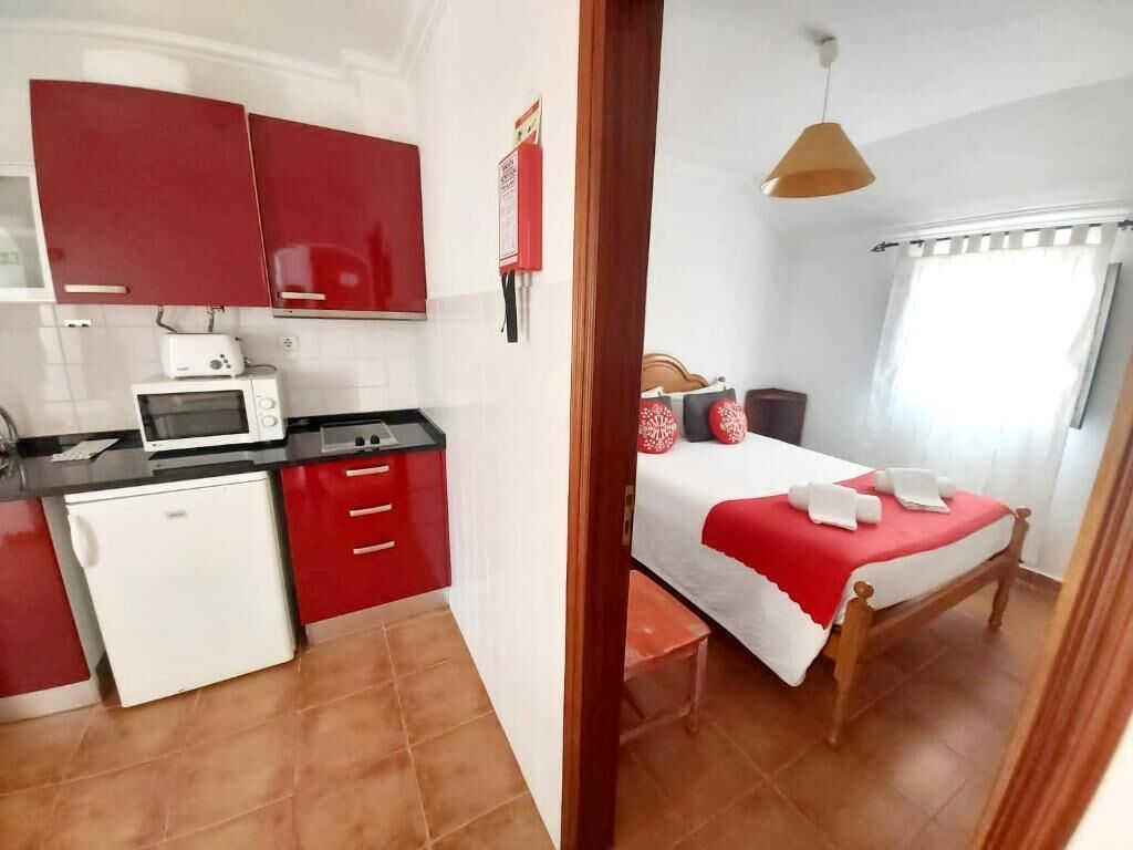 A compact room with a kitchenette, a bed with red bedding, and a window with curtains.