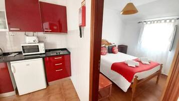 Habitación pequeña con kitchenette, una cama con ropa de cama roja y una ventana con cortinas.