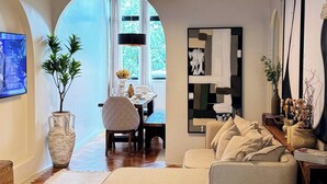 A modern living room with a herringbone wood floor, a large sofa, a dining area with natural light, and a ceiling with exposed beams. - Ipanema Premium Design · Renovated · Steps to Beach & Subway (Rio de Janeiro)