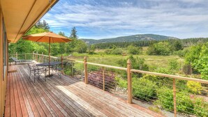 A wooden deck with a table and chairs, an umbrella, and a view of a green landscape with hills. - AUTUMN SALE! Cedar Field House on 2 Acres - horseshoes, Volleyball, Fire pit (Eastsound)
