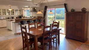 A modern kitchen with a central island, wooden dining table, and chairs. There are pendant lights, a large window with a view, and a wooden cabinet. - AUTUMN SALE! Cedar Field House on 2 Acres - horseshoes, Volleyball, Fire pit (Eastsound)