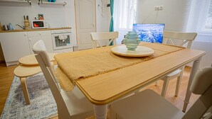 A compact kitchen with white cabinets, a microwave, and a small oven. There is a dining table with a beige placemat and white chairs. A television is mounted on the wall, and a window with curtains is visible. - Boutique flat in Akacfa street (Budapest)