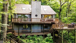 Een houten huis met twee verdiepingen, met een balkon op de bovenverdieping, omgeven door een dicht bos.