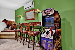Ein Zimmer mit grüner Wand, einer Wandklimaanlage, einer Bärenstatue, einem Tisch mit Stühlen und einer Videospielmaschine.
