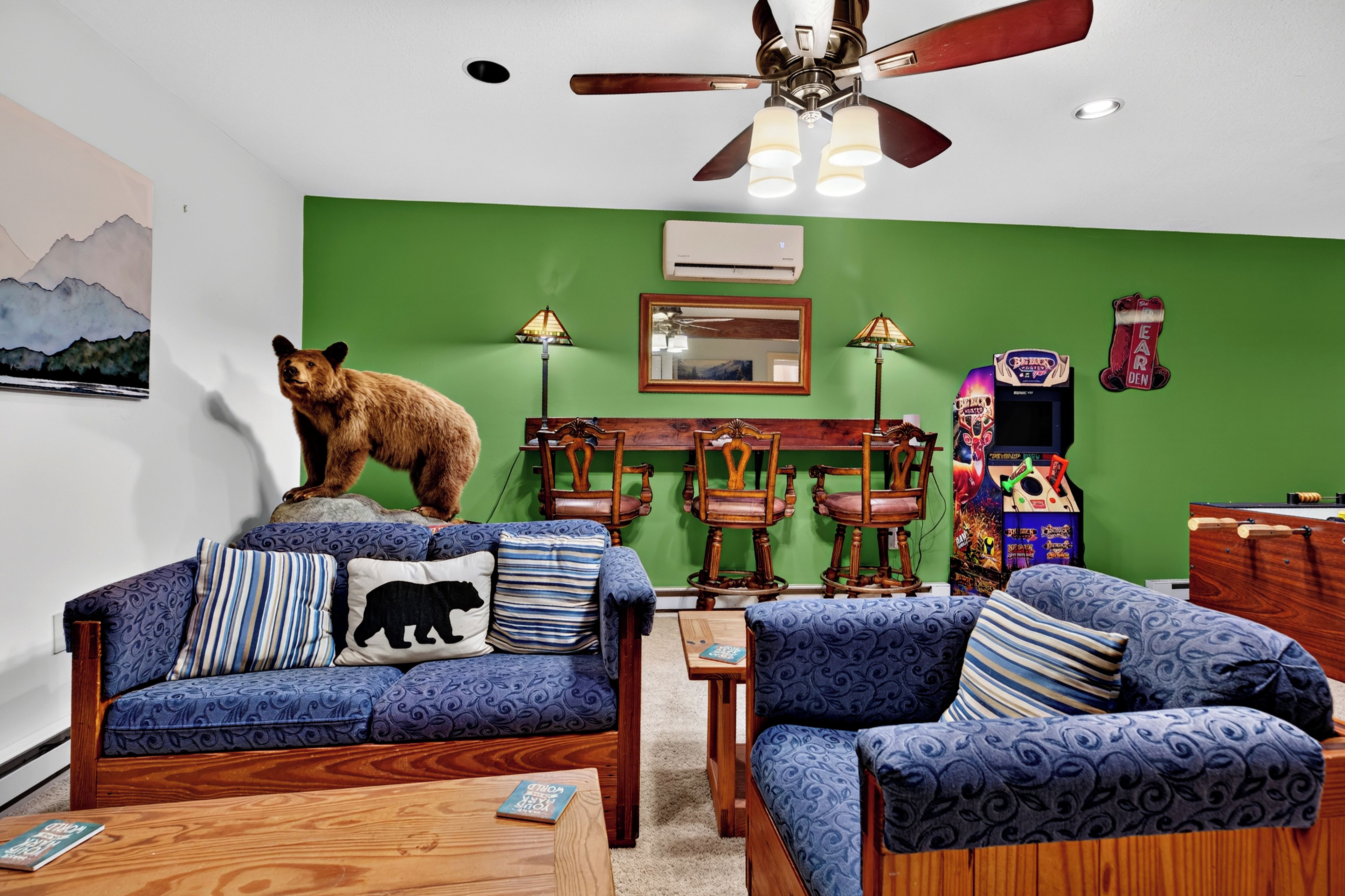 A room with a green wall, a ceiling fan, a sofa with patterned cushions, a chair, a small table, and a bear decoration.