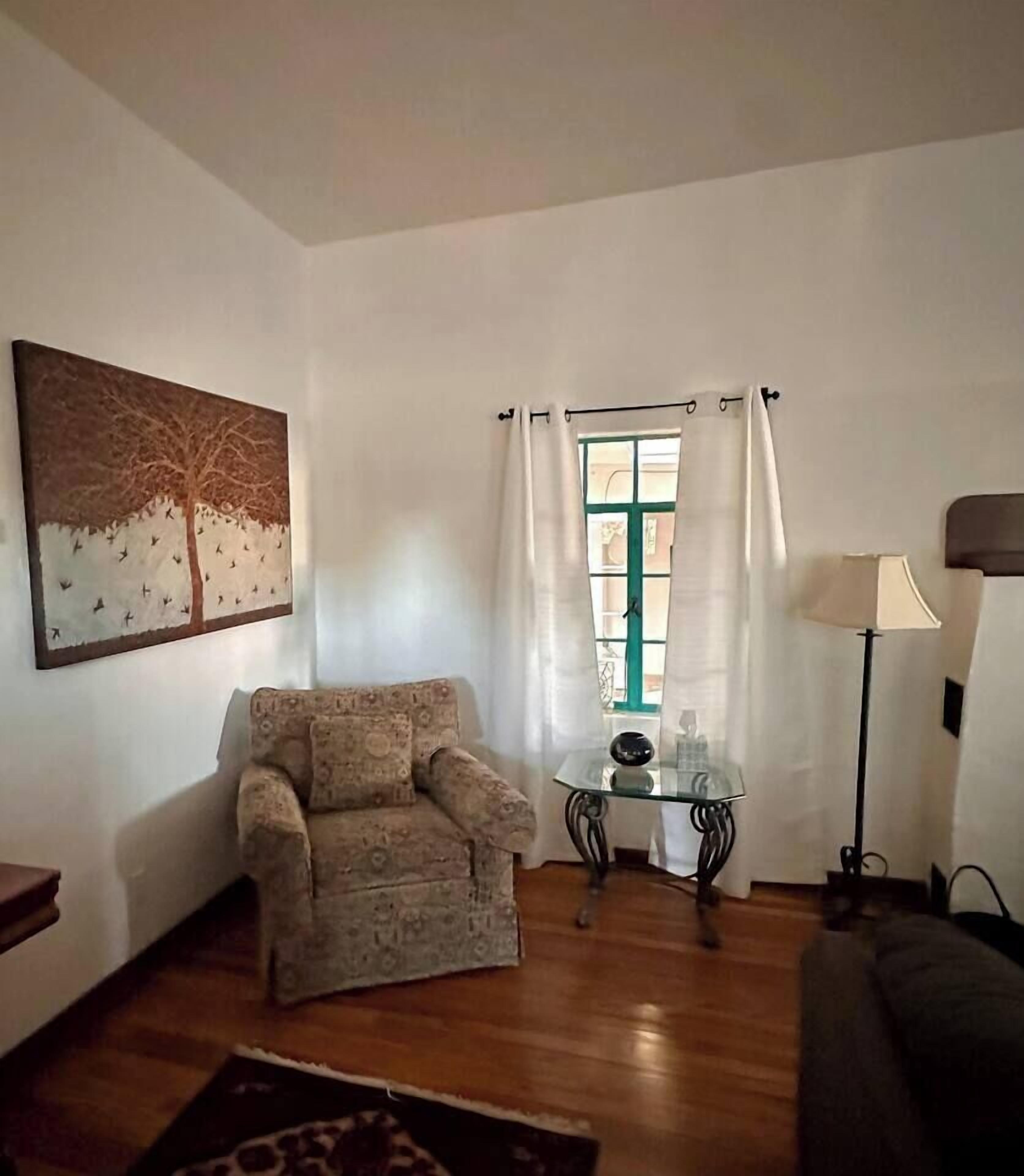 Ein Wohnzimmer mit einem gemusterten Sessel, einem Glastisch mit einem dekorativen Gegenstand, einer Lampe und einem Baum-Motiv mit Vögeln.