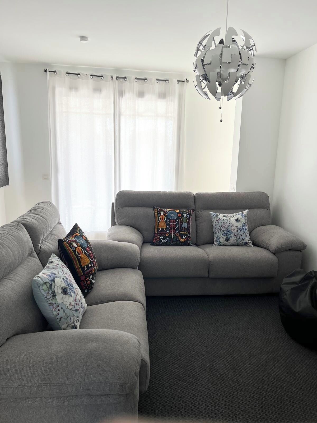 A modern living room with a grey sofa set, patterned cushions, a hanging metallic chandelier, and sheer curtains.