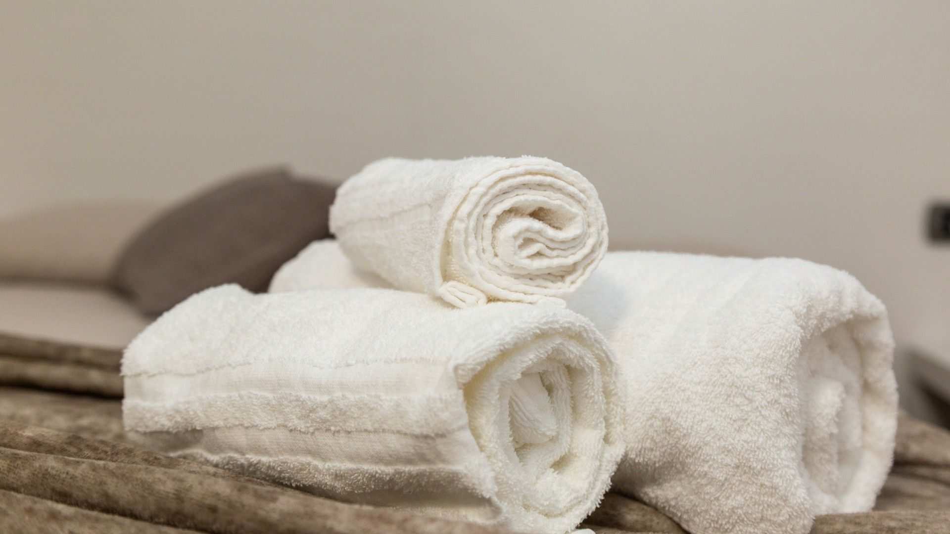 A neatly folded white towel on a textured surface, possibly a bed or couch, with a neutral-toned pillow in the background.