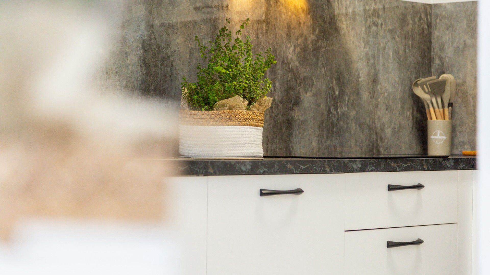 A kitchen with a plant in a woven basket, a utensil holder, and a marble countertop.