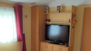 Una stanza con un mobile TV in legno, un televisore a schermo piatto, un tavolino in legno, un letto con coperta rosa e una finestra con tende.