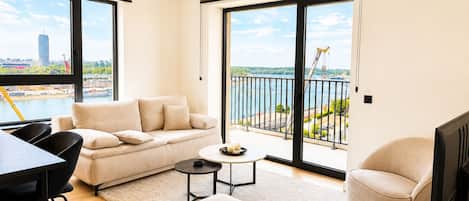 Una sala moderna con sofá, reposapiés y sillones, una mesa de comedor y un balcón con vista a la ciudad y al agua.