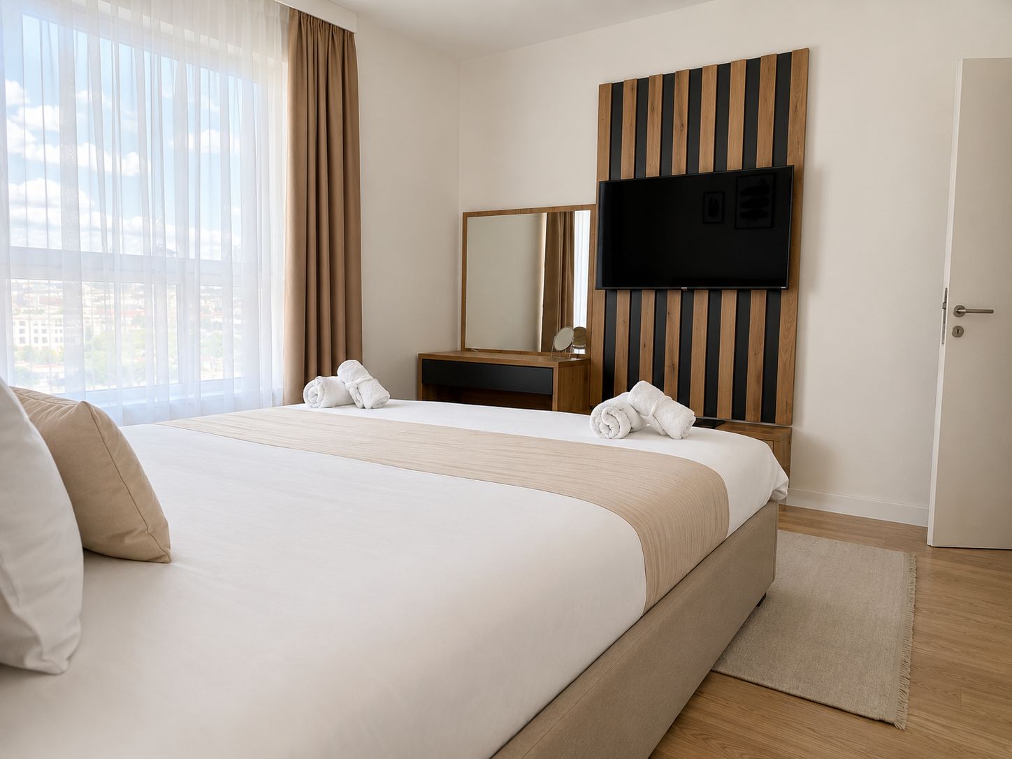 A hotel room with a large bed, a flat-screen TV mounted on a wooden panel, a bedside table with a mirror, and a view of the city through large windows.
