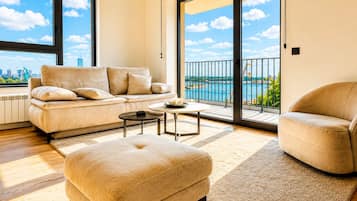 Una sala moderna con sofá, reposapiés y sillón, una mesa redonda y vistas a un cuerpo de agua a través de amplias ventanas.
