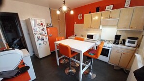 Een kleine keuken met een witte koelkast, oranje stoelen, een witte tafel en houten keukenkastjes.