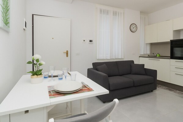 Un salon moderne avec une table à manger blanche, un canapé gris foncé et un coin cuisine doté de meubles blancs et d’un four noir.
