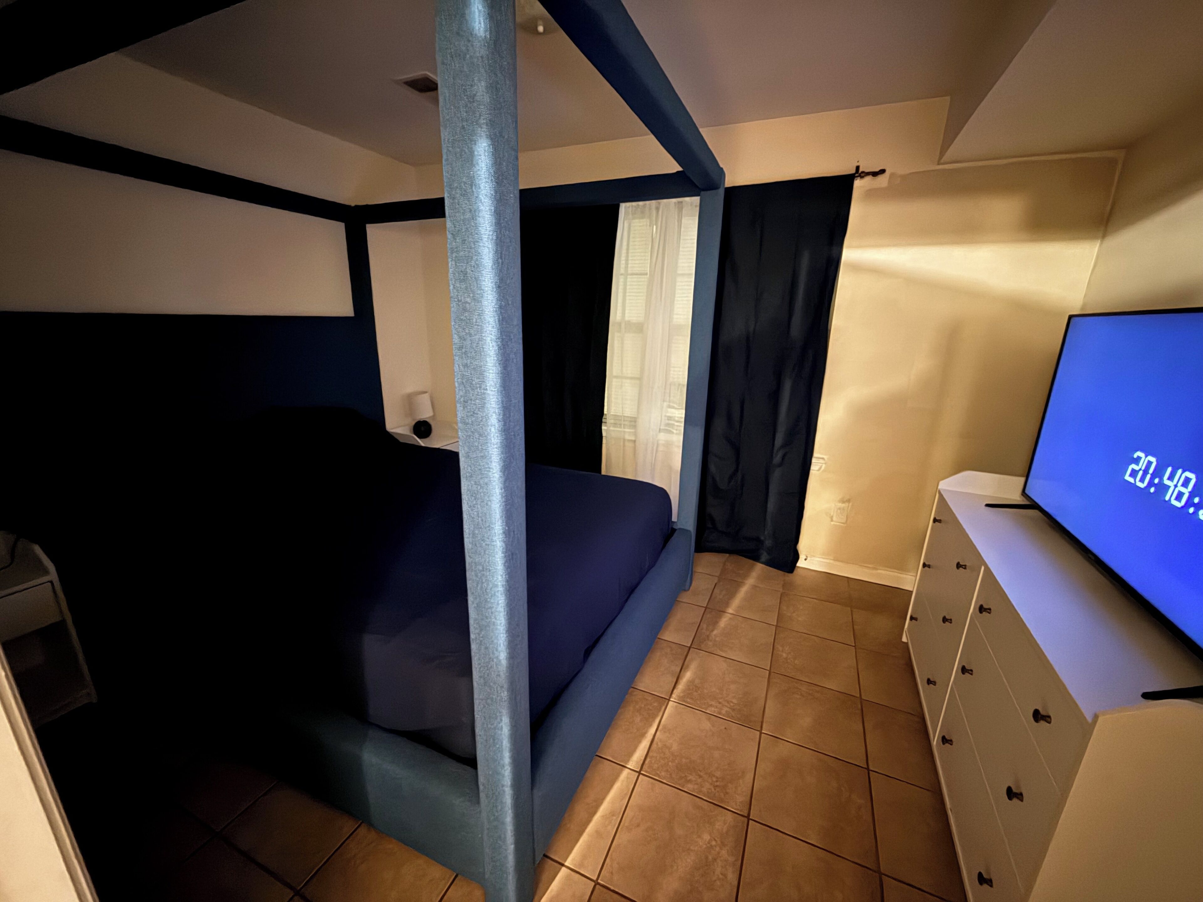 Une petite pièce comprenant un lit, une commode, un téléviseur affichant l’heure et une fenêtre avec des rideaux.