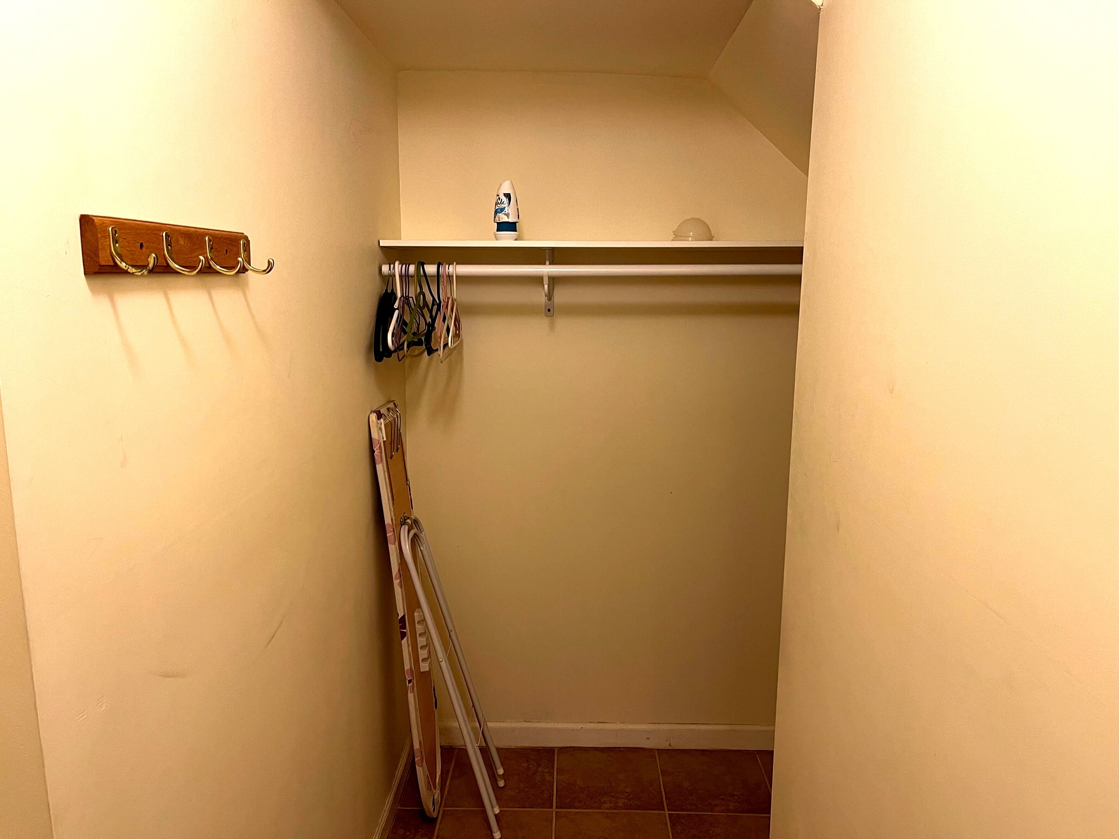 A small, narrow hallway with a wooden coat rack, a set of crutches leaning against the wall, and a white shelf with a bottle on top.