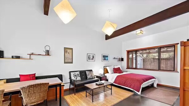 Quarto moderno com mesa de jantar de madeira, sofá, cama com cobertor vermelho e uma janela grande.
