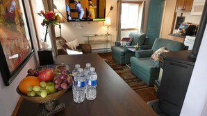 Een woonkamer met een houten tafel, een schaal fruit, waterflesjes, een televisie, een bank en een keukenhoek.